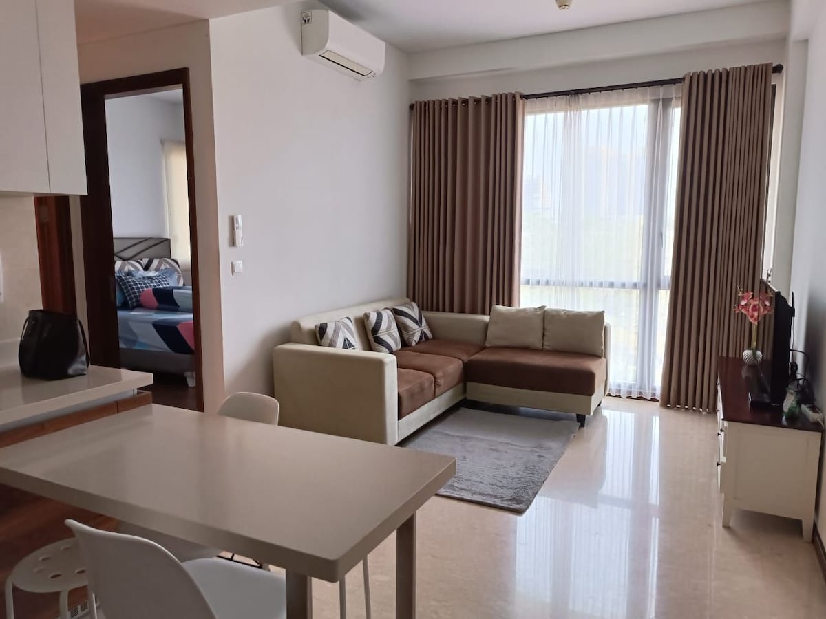 A light-filled living area features a comfortable sectional sofa facing a flat-screen television. A dining table with white chairs is positioned nearby. Large windows are adorned with sheer curtains, allowing natural light to illuminate the space, while beige and brown tones create a neutral palette.