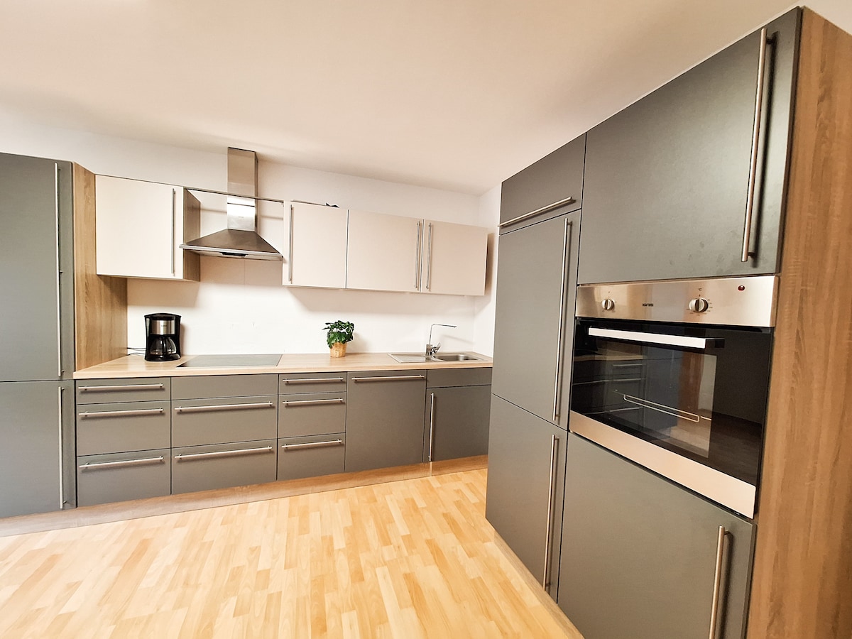 The kitchen features modern cabinetry with a mix of dark and light tones. Stainless steel appliances, including an oven and coffee maker, are integrated into the design. A potted plant adds a touch of greenery, while the spacious layout provides ample room for meal preparation.