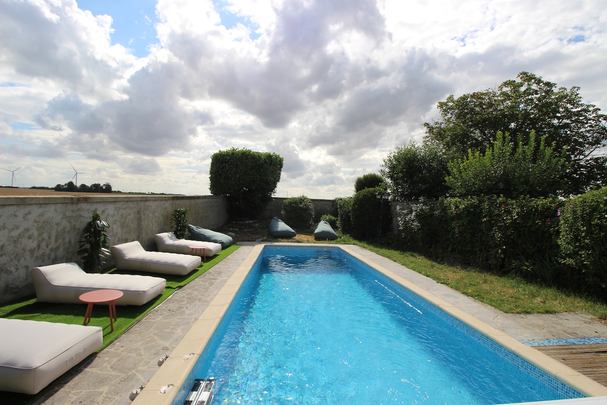 A tranquil outdoor pool area is presented, featuring a rectangular swimming pool surrounded by lush greenery. Comfortable lounge chairs are arranged along the poolside, with soft cushions for relaxation. A small round table is placed next to the pool, enhancing the serene atmosphere.