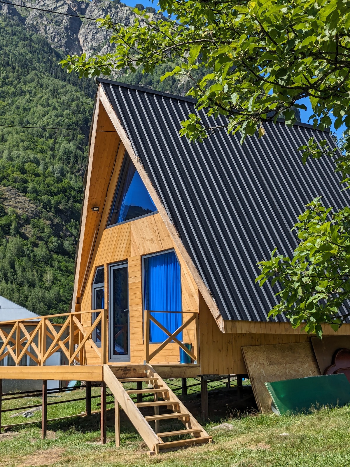 The cottage's exterior features a distinctive A-frame design with wooden paneling and a sloped roof. Large glass windows provide ample natural light, while a wooden deck with railings offers a welcoming entrance. Lush greenery and mountainous terrain surround the structure, contributing to a serene environment.