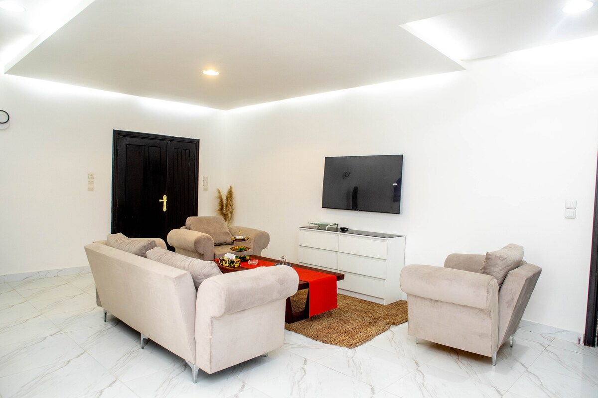 A spacious living area features a pair of light-colored sofas arranged around a central coffee table. A modern television sits on a low, white cabinet against the wall. Natural light fills the room, complemented by soft, neutral tones and sleek flooring.