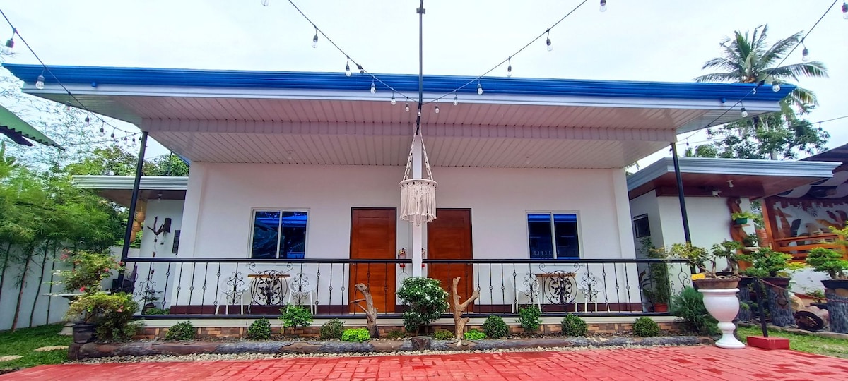 The exterior features a one-story house with a blue roof and a white facade. A covered porch with a railing is adorned with decorative plants. Two wooden doors and several windows allow natural light to enter, while string lights add charm to the outdoor space.