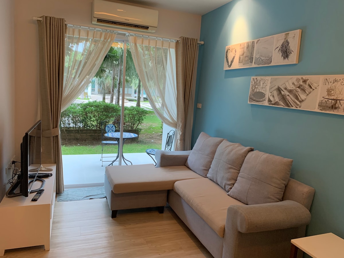 A cozy living area features a light grey sectional sofa, positioned near a television. Sheer curtains frame large windows, allowing natural light to enter. A small outdoor seating area with a round table and two chairs is visible beyond the glass doors, surrounded by greenery.