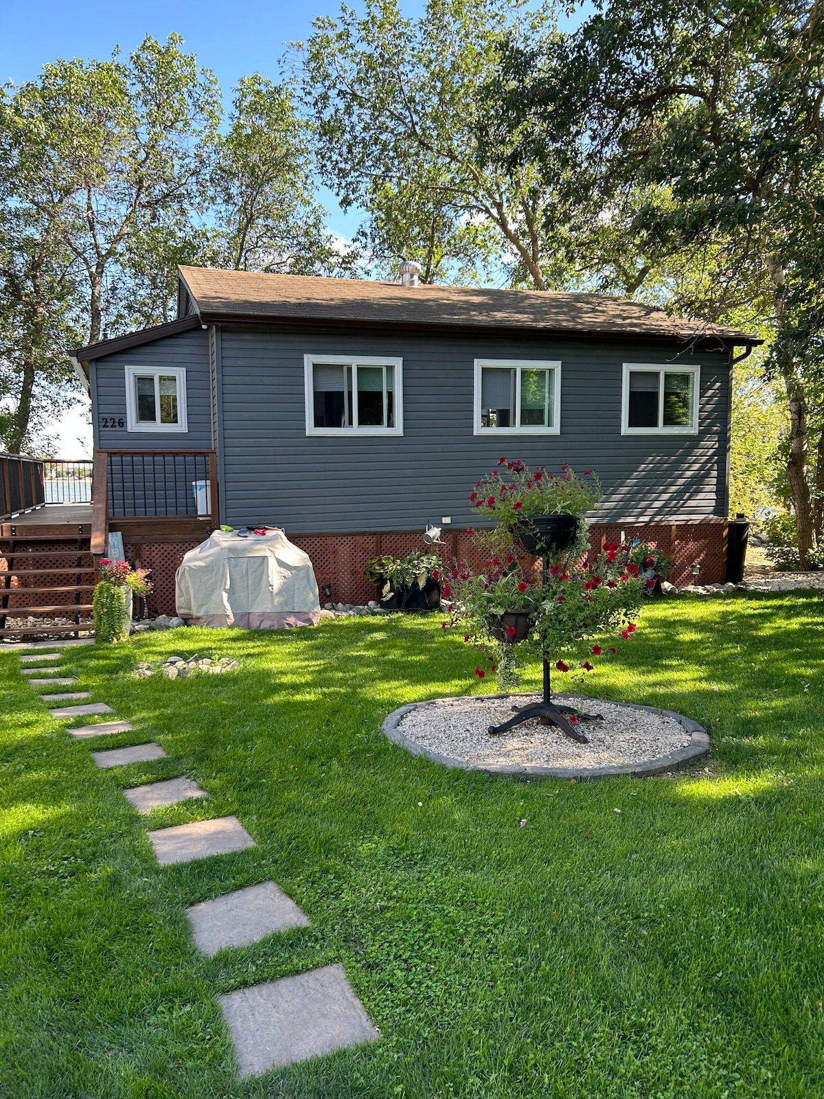 A well-maintained home with grey siding is set within a green lawn. A short pathway of stepping stones leads to the entrance, while a BBQ grill is visible on the deck. Surrounding trees provide shade, and flower beds add color to the outdoor space.