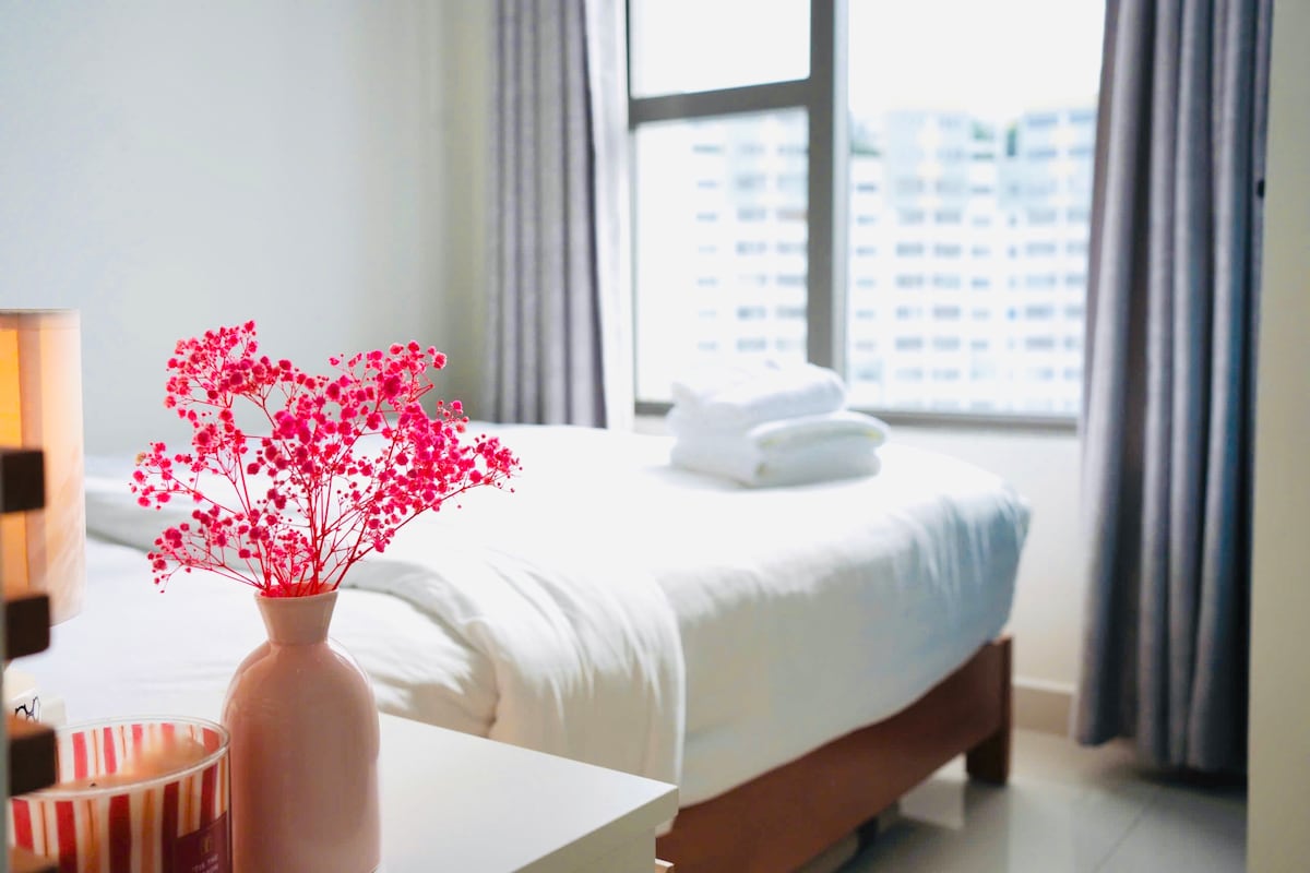 A spacious bed is neatly made, with fresh white linens and towels stacked at the head. A small table holds a pink vase filled with decorative flowers, alongside a candle. Large windows with gray curtains allow natural light to fill the room.