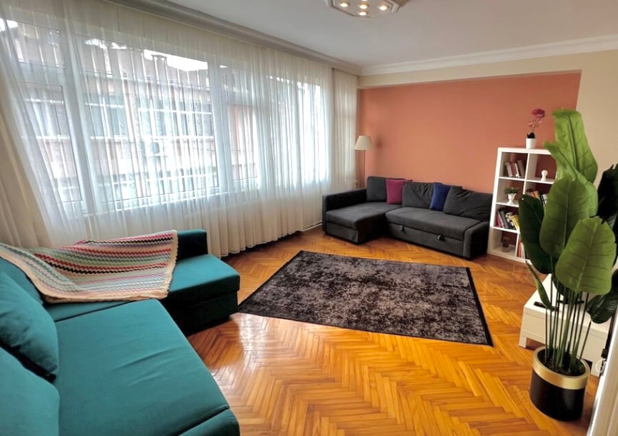 A spacious living area features a plush dark rug on a polished wooden floor. A corner sofa in charcoal gray is complemented by colorful cushions, while a cozy teal sofa is adorned with a knitted blanket. Large windows allow ample natural light to fill the space.