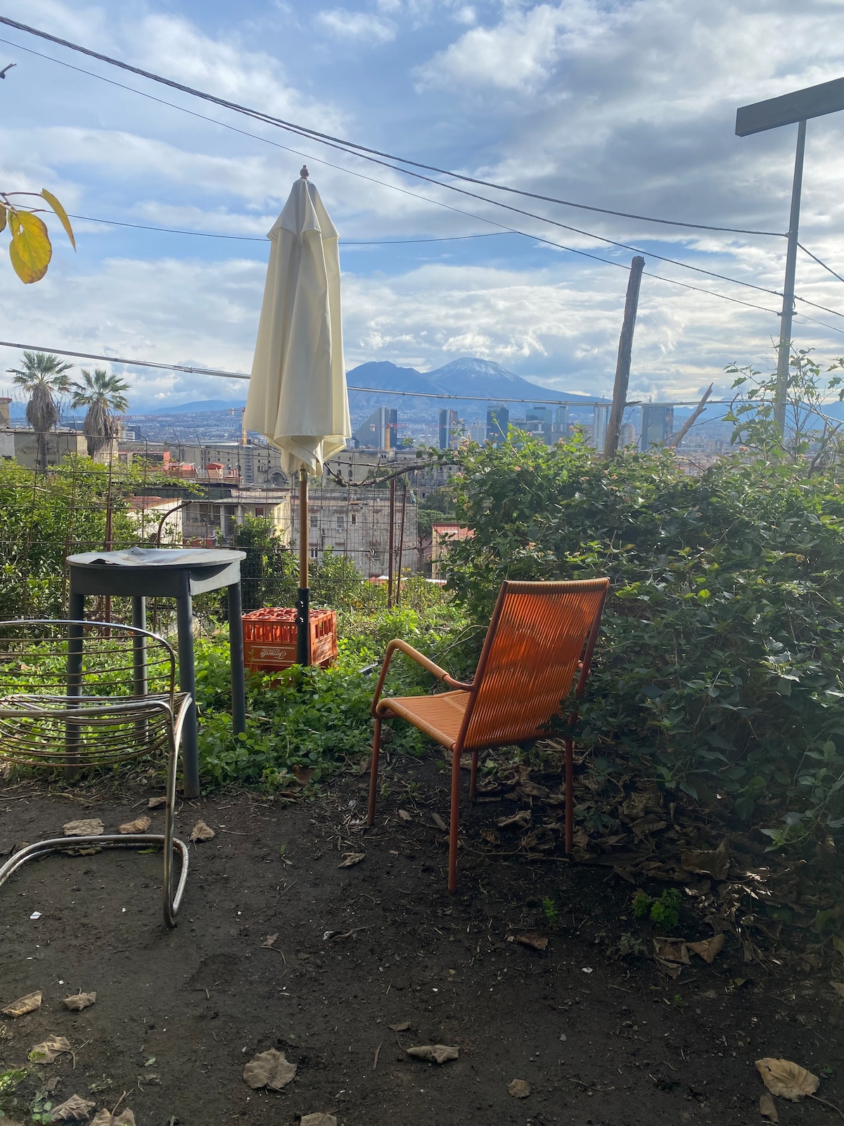 A small outdoor seating area features an orange chair and a cafe table, surrounded by greenery and a view of distant mountains. A white umbrella stands to the side, providing shade. The area indicates a peaceful spot for relaxation.
