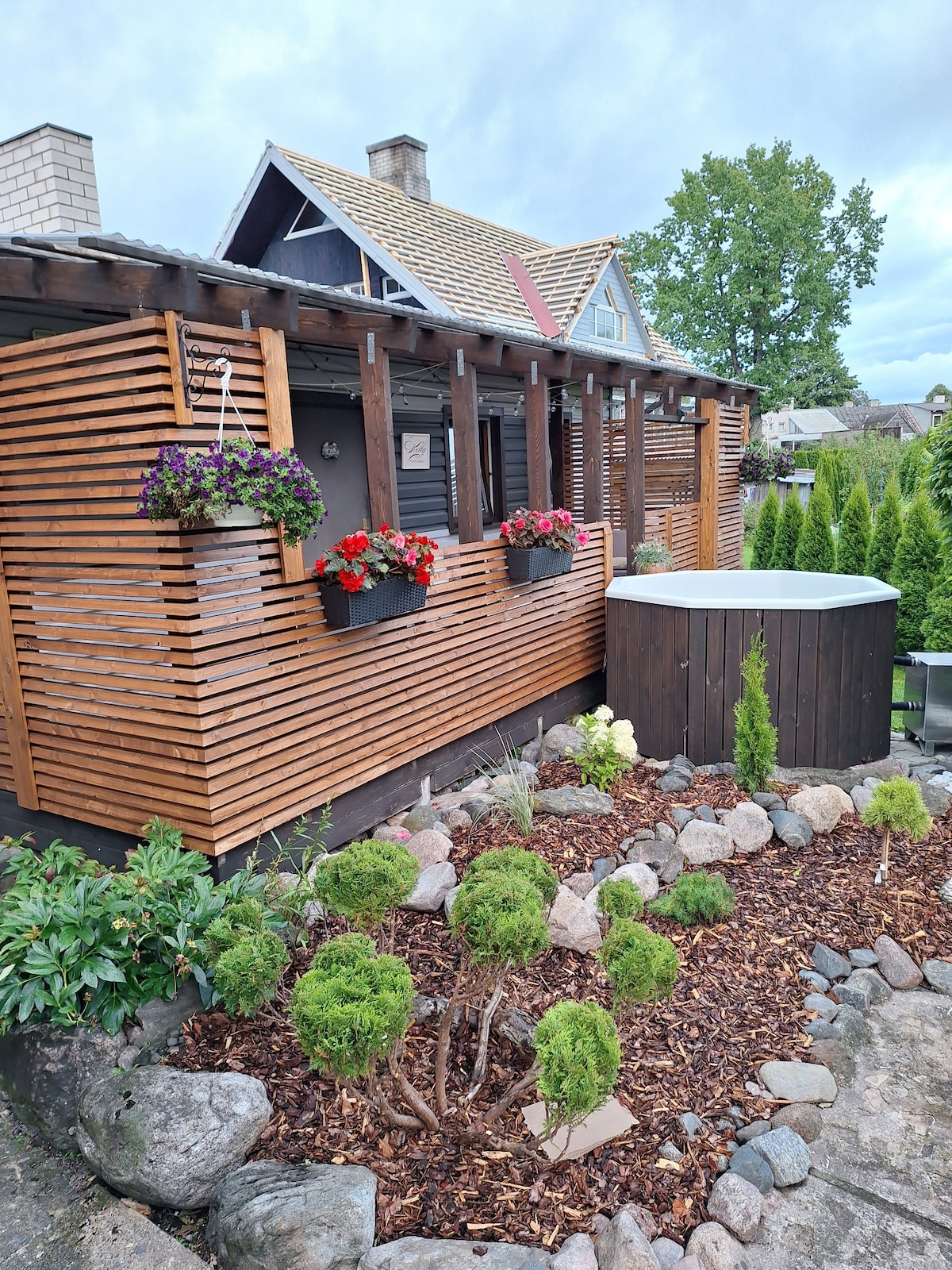 The exterior of the sauna house is framed by wooden slats and decorated with vibrant flower boxes. A neatly arranged garden bed complements the structure, featuring small shrubs and stones. A hot tub is positioned to the side, enhancing the garden's tranquil setting.