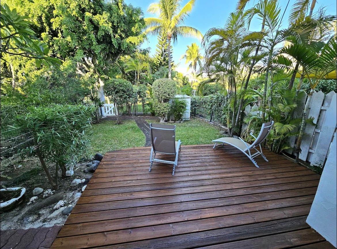 A private wooden deck is surrounded by lush greenery, featuring two lounge chairs positioned for relaxation. Tropical plants and palm trees create a tranquil atmosphere, with a grassy area visible in the background. The space offers a sense of seclusion and comfort.