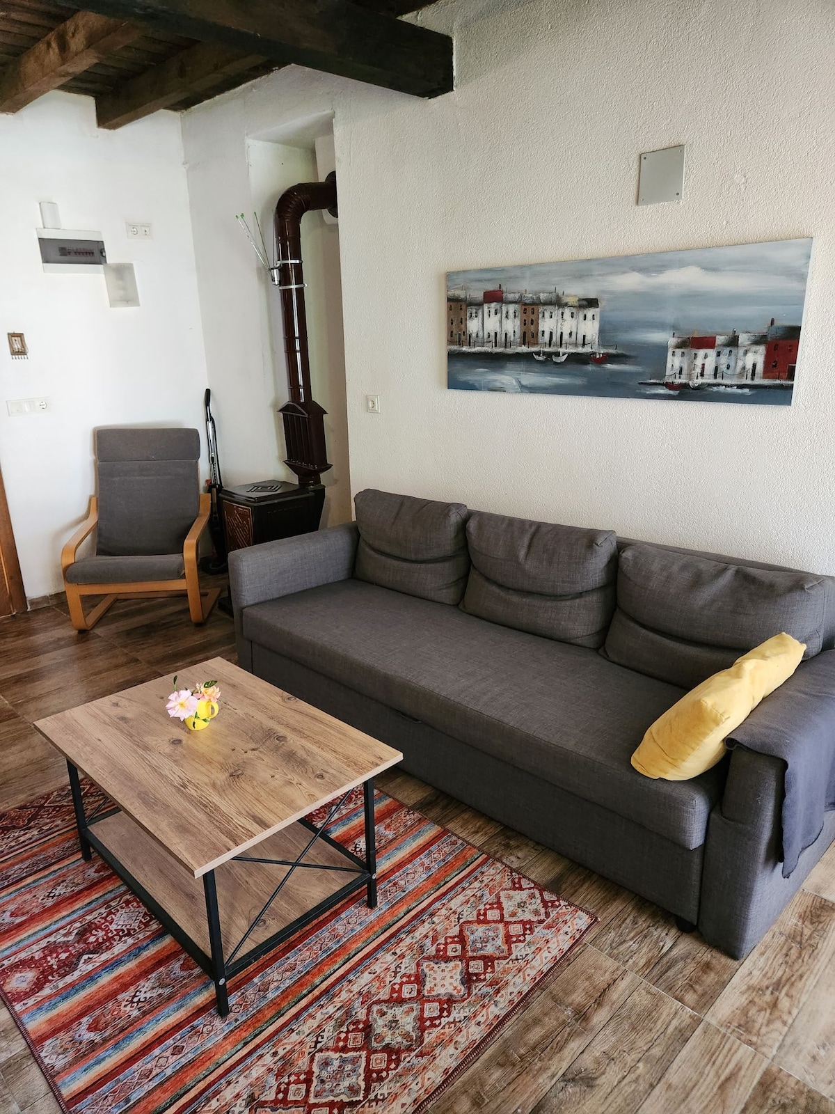 A welcoming living area features a grey sofa complemented by a yellow cushion and a wooden coffee table. A decorative rug adds warmth to the floor. A painting depicting a coastal scene decorates the wall, while a chair is positioned nearby, creating a cozy reading nook.