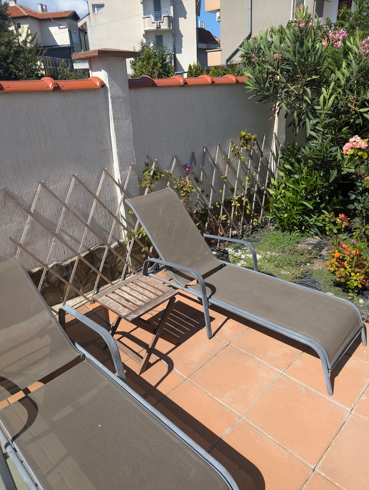 Two lounge chairs are positioned on a tiled patio, accompanied by a small side table. Surrounding vegetation includes a variety of flowering plants and greenery, contributing to a serene outdoor space. A wooden trellis is visible along one boundary, enhancing the garden's charm.