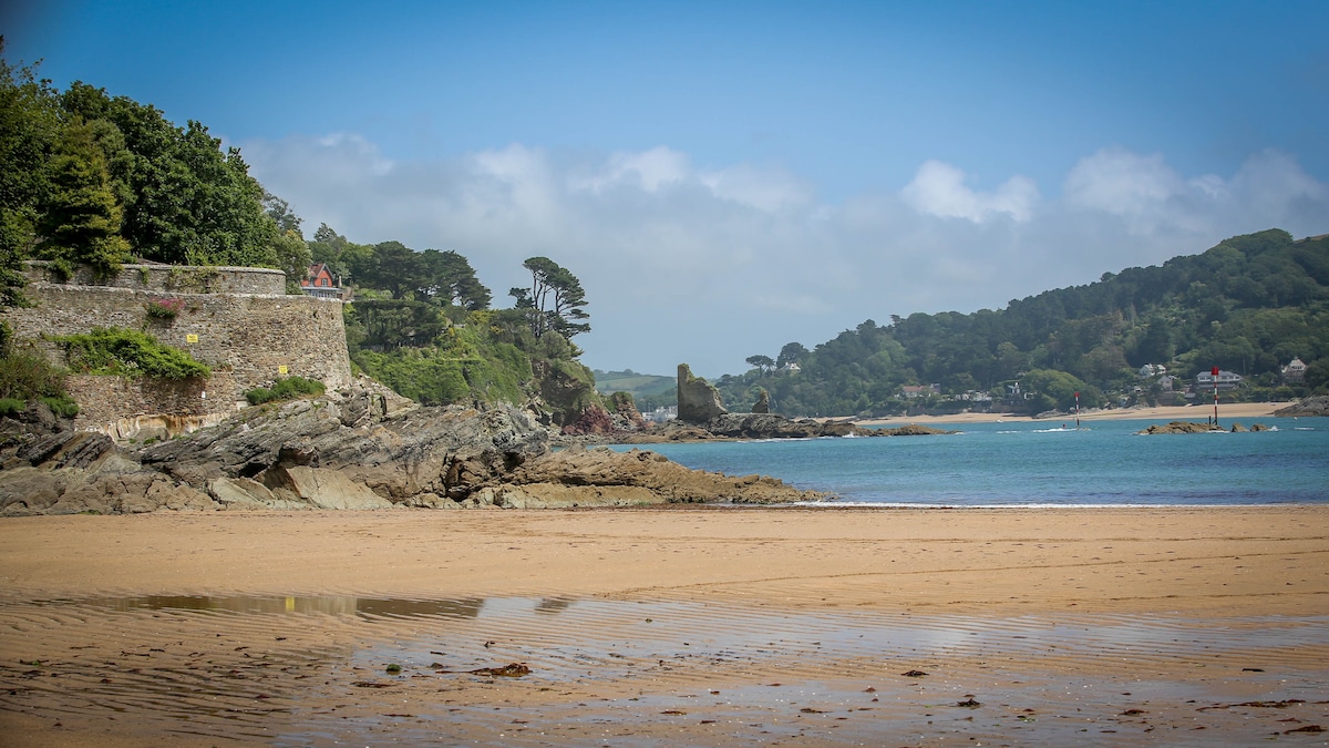 A sandy beach stretches along a calm, blue sea, framed by rocky outcrops and lush green hills. A historic stone wall and trees are visible in the background, adding to the natural scenery. Sunlight glints off the water, enhancing the tranquil atmosphere.