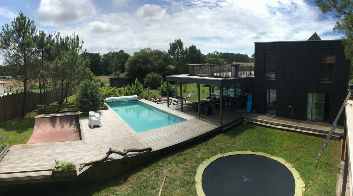 An outdoor view of the villa showcases a wooden deck surrounding a rectangular pool. Nearby, a trampoline and a small skate ramp are visible, along with lounge chairs. Lush greenery and trees create a natural backdrop, enhancing the serene atmosphere of the outdoor space.