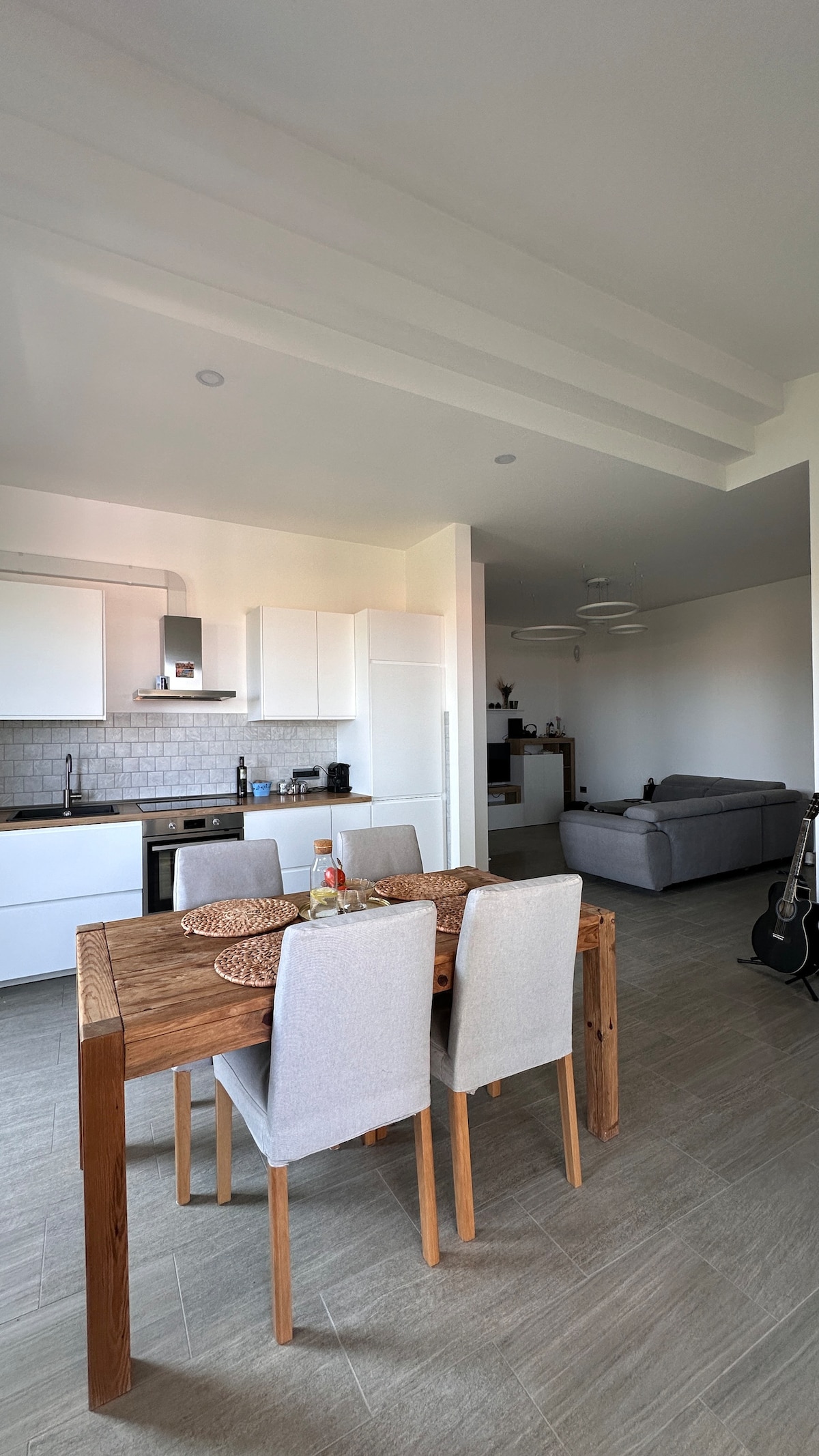 A modern kitchen and dining area feature a wooden table set with four light-colored chairs. The open-space layout includes sleek white cabinetry and stainless-steel appliances. A gray sofa is visible in the background, enhancing the relaxed atmosphere of the room.