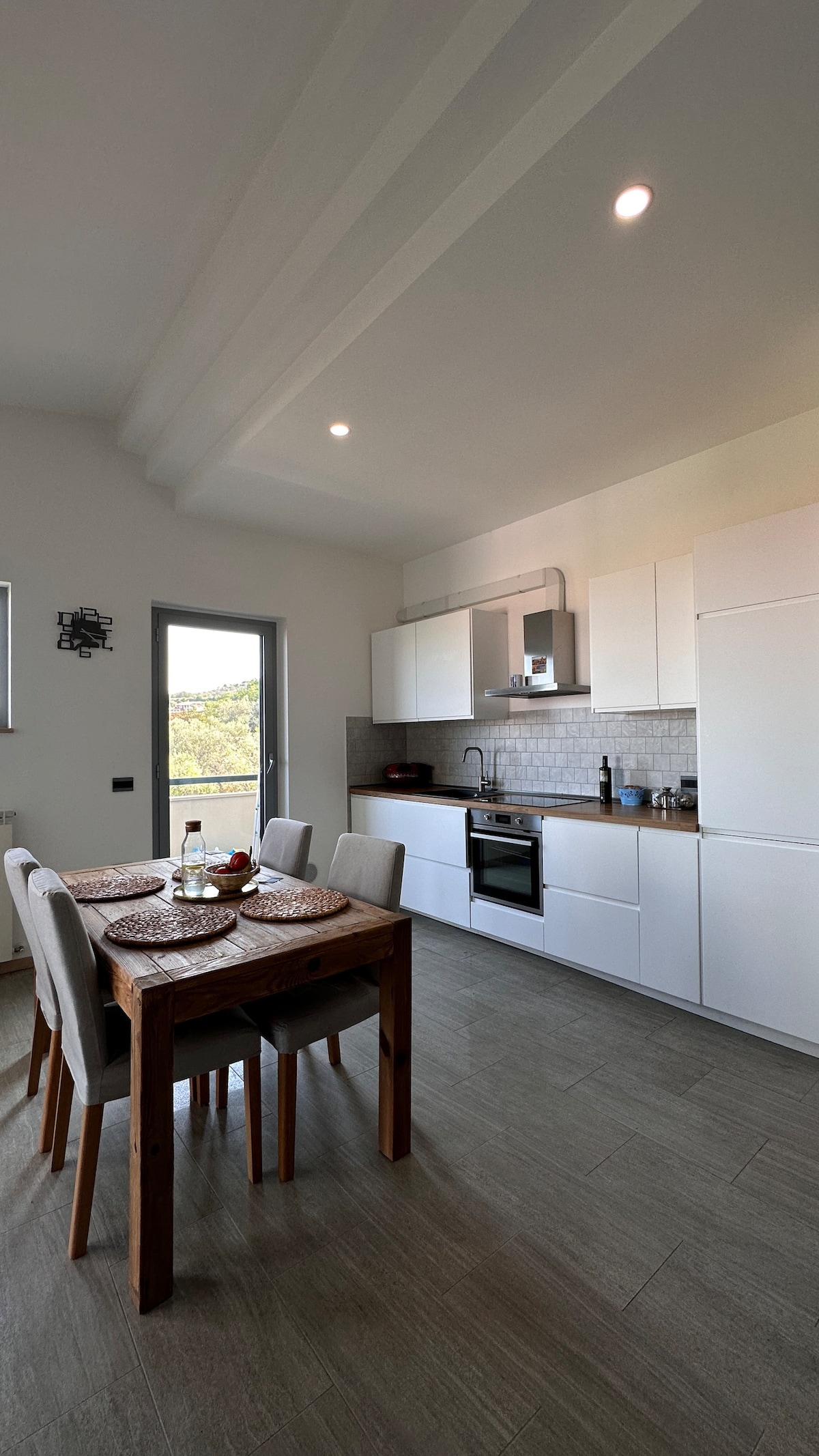 A modern kitchen is featured with sleek white cabinets and a contemporary stove. A wooden dining table is accompanied by four chairs, set with woven placemats. Natural light enters through a large window, providing a view of the outdoors.