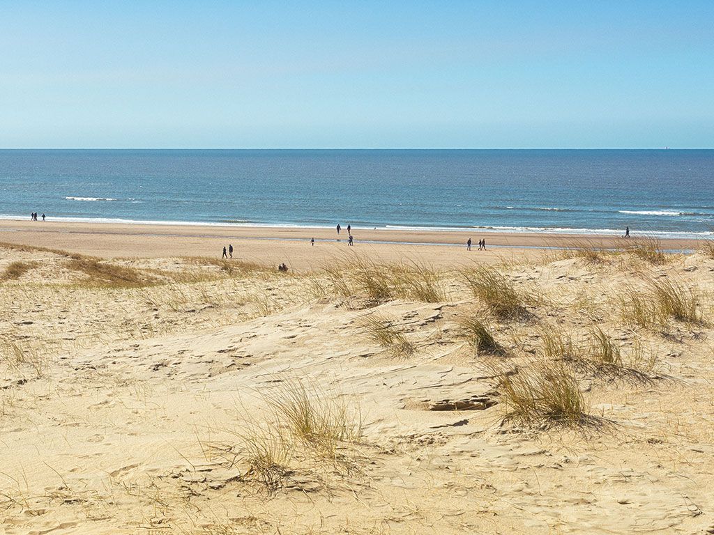 A sandy beach is visible, framed by natural dunes adorned with tufts of grass. The calm sea stretches into the distance, meeting a clear sky. Several figures can be seen walking along the shoreline, illustrating the beach's spaciousness.