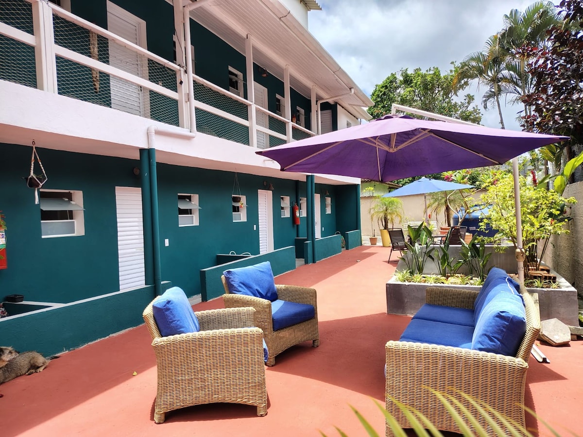 An outdoor seating area includes two wicker chairs with blue cushions positioned around a coffee table. A large purple umbrella provides shade, while green plants and a small dining set are visible in the background against a bright red floor.