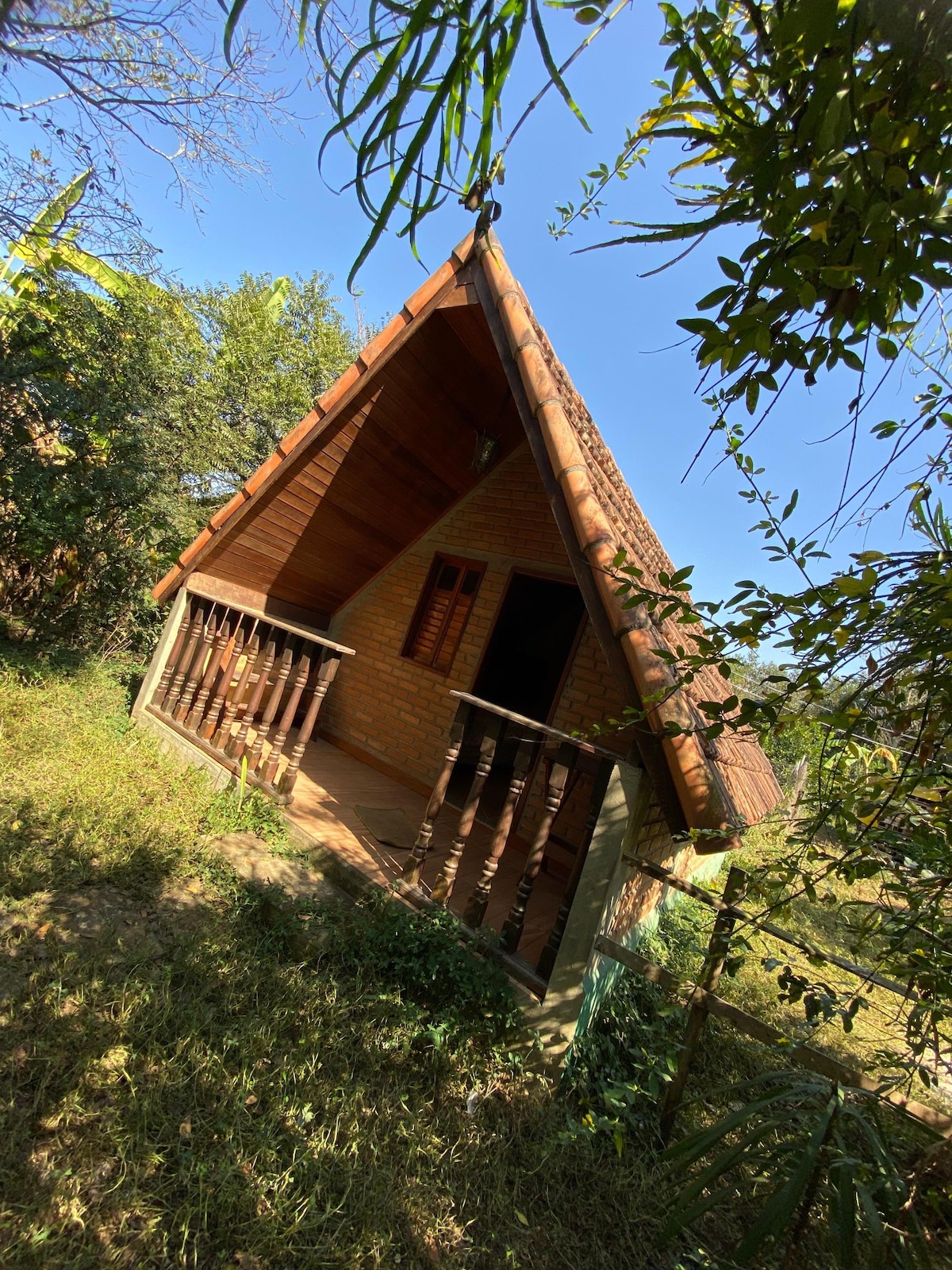 A charming A-frame cabin presents natural textures, with wooden accents and a terracotta roof under a clear blue sky. Surrounded by trees and greenery, a spacious porch with wooden railings invites relaxation in a serene outdoor setting.
