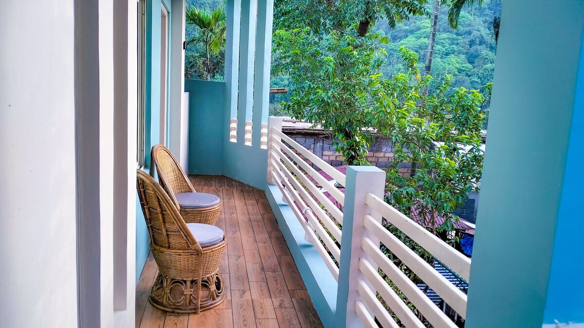 A spacious balcony features two woven chairs and a small table, set against a backdrop of lush greenery. Natural light filters through the railings, creating a serene outdoor space for relaxation.