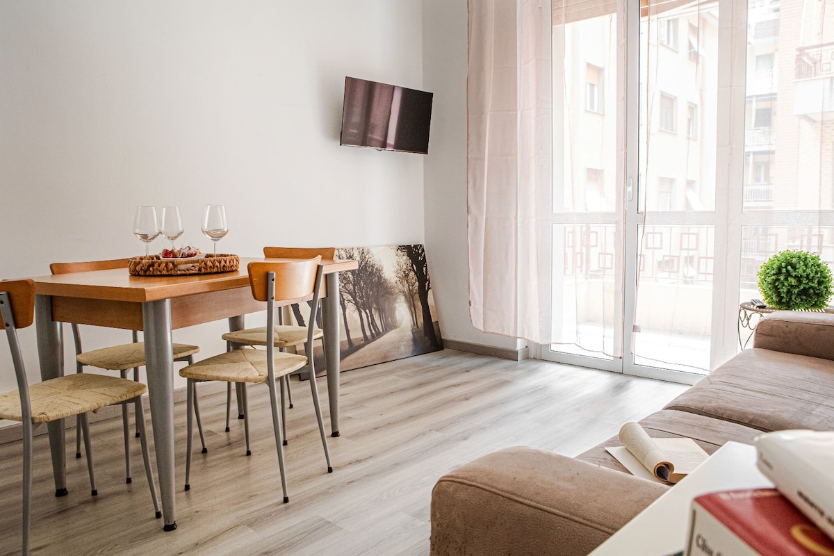 A dining area features a wooden table surrounded by four chairs. A small television is mounted on the wall, and natural light enters through large glass doors. A cozy sofa is positioned to create an inviting atmosphere, with a decorative plant adding a touch of greenery.