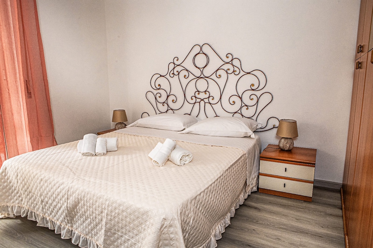 A spacious bedroom features a decorative headboard above a large bed with crisp linens. Two bedside tables with lamps are positioned on either side, and light flows in through sheer curtains, creating a bright and airy space.
