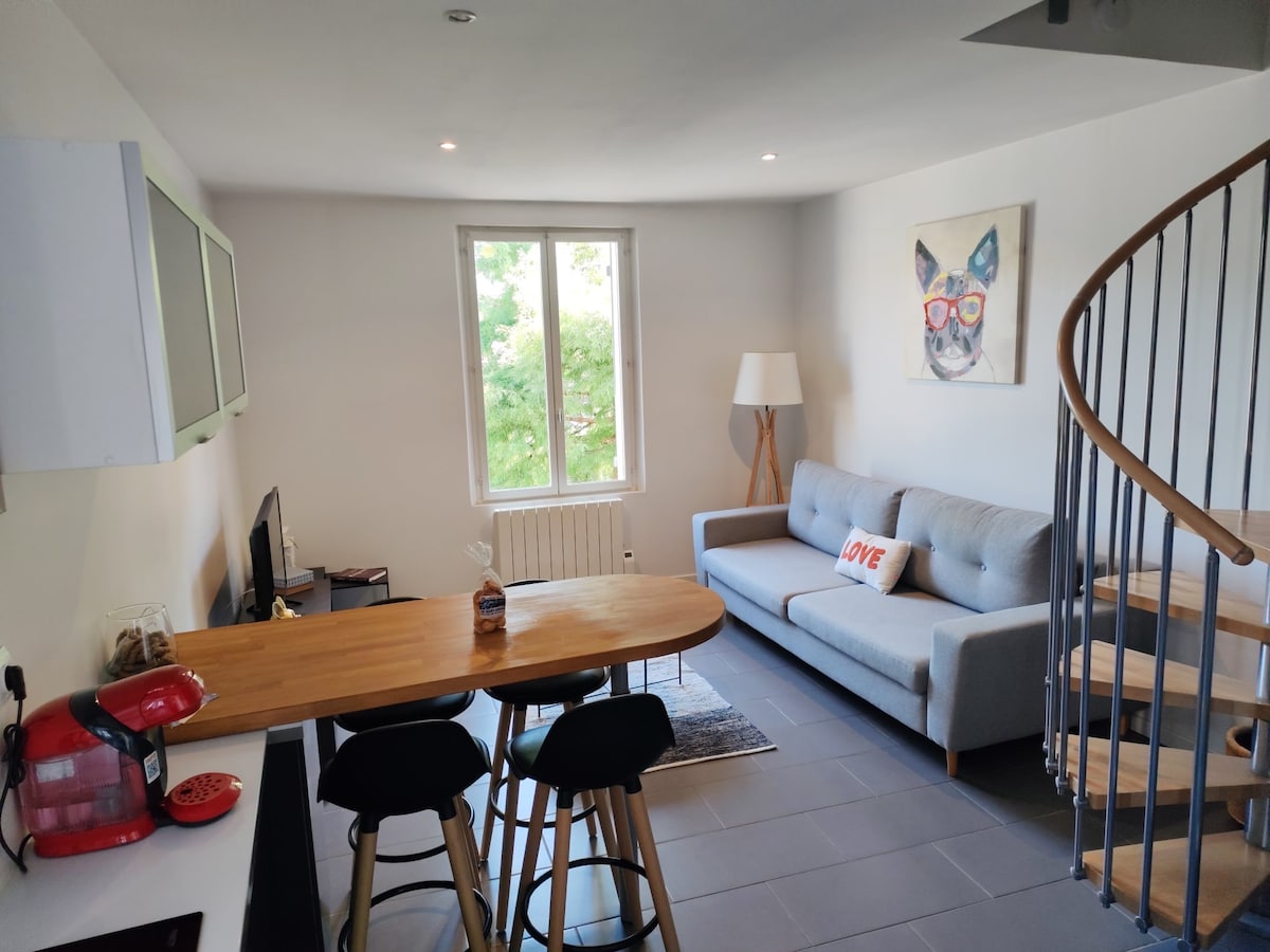 A bright living area features a gray sofa beside a large window, allowing natural light to fill the space. A wooden table with black stools is positioned nearby, and a spiral staircase leads to an upper level. A playful wall art piece adds character.