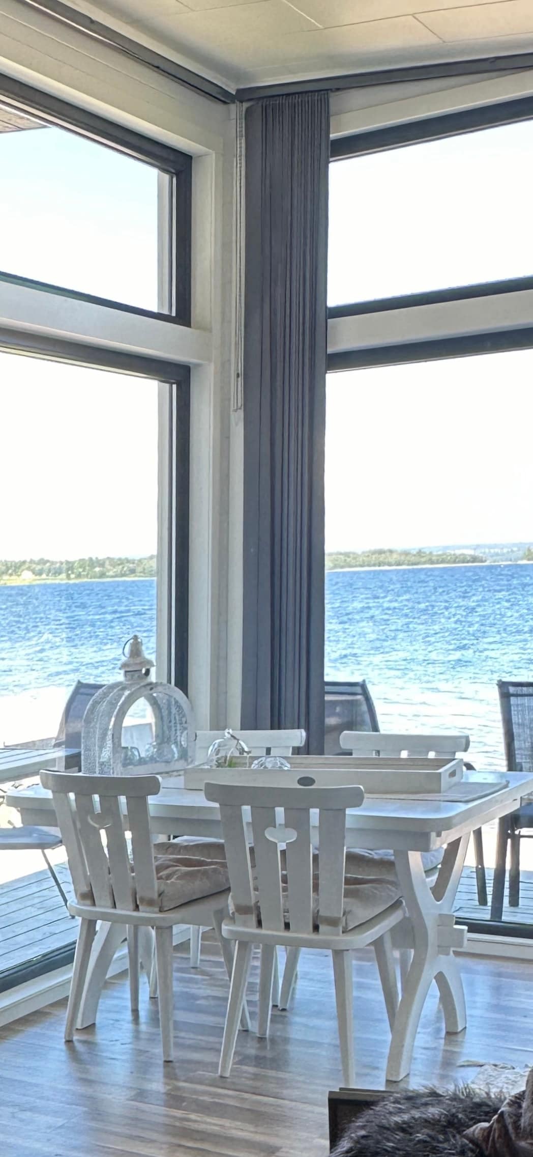 A dining area is showcased with a white table surrounded by four matching chairs, set against large windows. The views of the expansive water with gentle waves are visible, enhancing the serene atmosphere of the space.