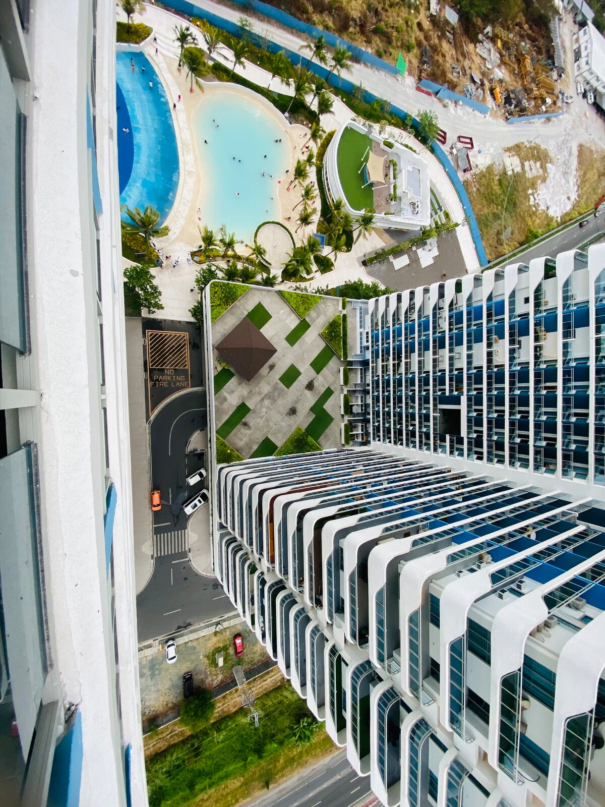 The image presents a bird's-eye view of a carefully designed complex. Lush palm trees surround the pool area below, complemented by landscaped green spaces. The modern architecture of the building features white and blue accents, showcasing a harmonious blend with the serene environment.