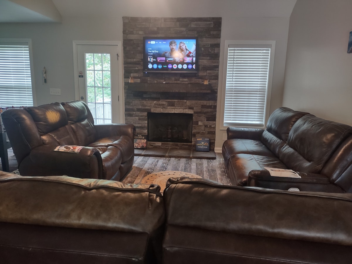 A spacious living area showcases two luxurious leather sofas arranged around a stone fireplace. A modern flat-screen TV is mounted above the hearth, providing entertainment options. Natural light filters in through the windows, creating an inviting atmosphere for relaxation and socializing.