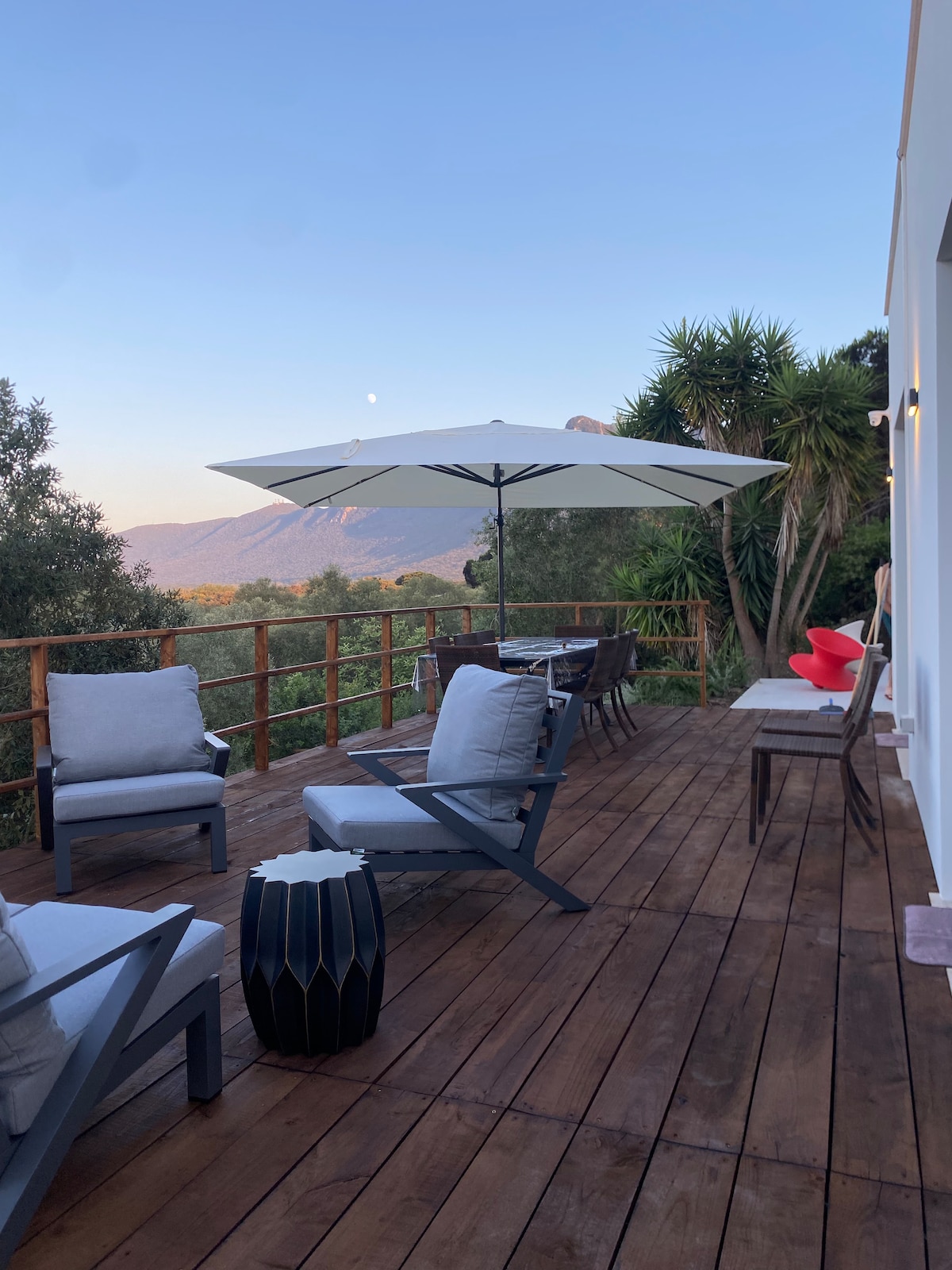An outdoor living space is seen, featuring a wooden deck with comfortable seating. A large umbrella provides shade over a dining area, accompanied by a small black accent table. Lush greenery and distant mountains create a serene backdrop, with a gentle evening sky visible.