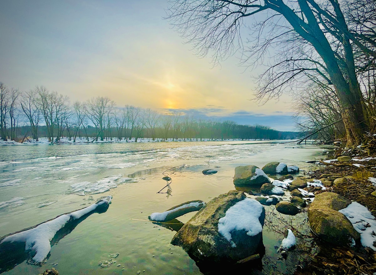 The tranquil scene captures the Grand River during winter, with icy banks and patches of snow on rocks. Bare trees line the river, reflecting the soft light of the setting sun against the icy water.