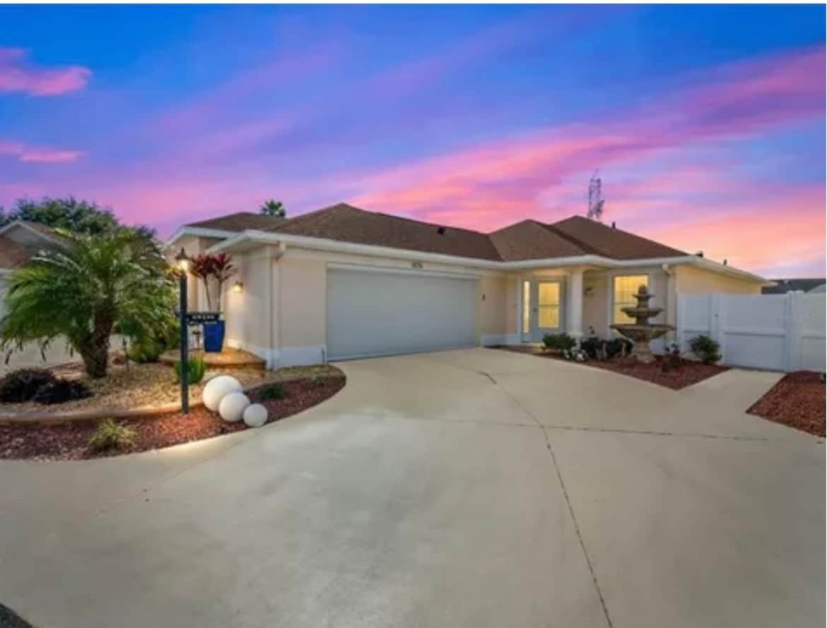 A welcoming exterior view of a single-story home is presented, featuring a spacious driveway and a well-maintained landscape. Decorative elements, such as palm trees and shrubbery, surround the entrance. The sky showcases soft hues of pink and blue during twilight, adding a peaceful ambiance.