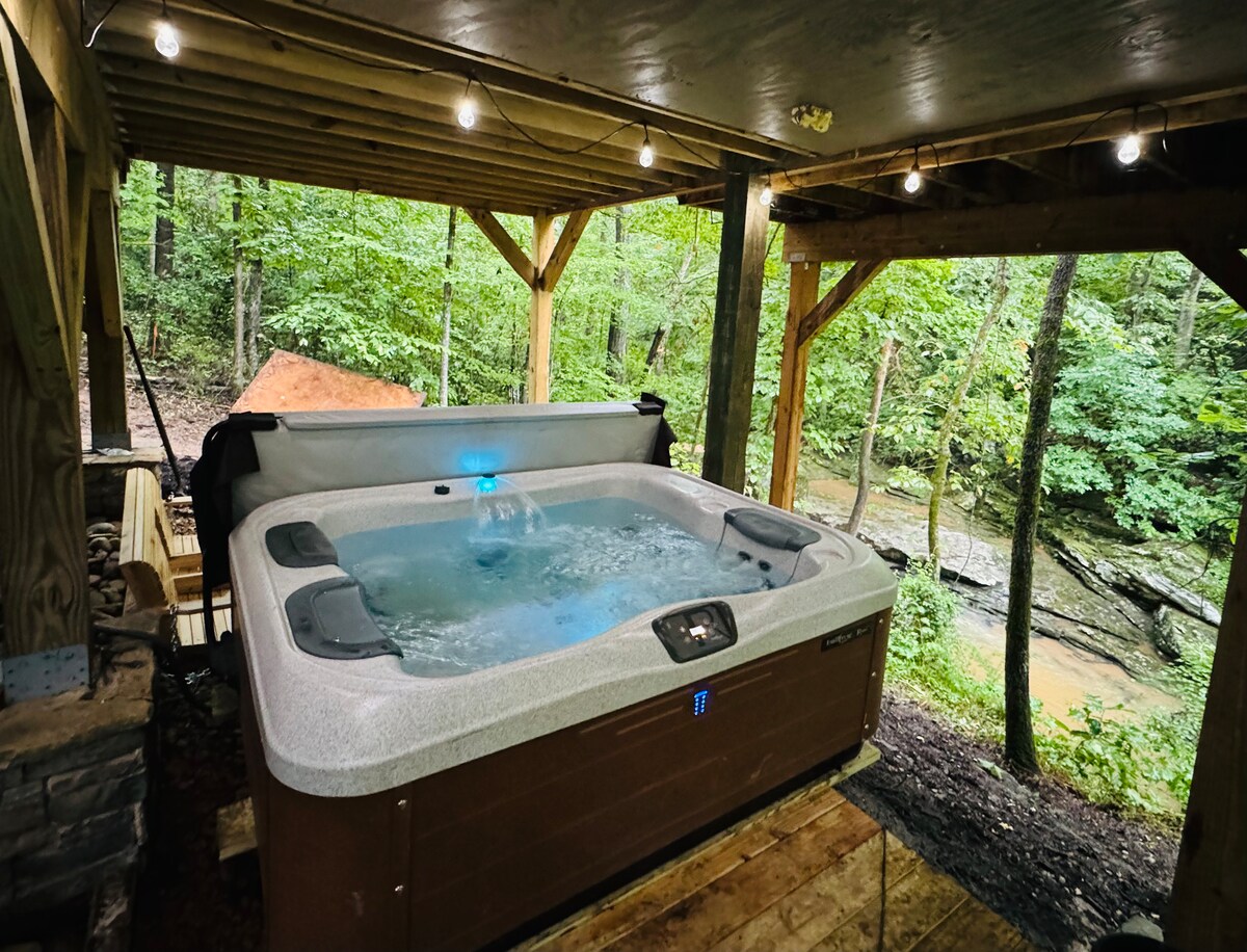 A hot tub is positioned under a wooden structure, surrounded by greenery. Soft lighting is strung above, creating a tranquil ambiance. The flowing creek is visible through the trees, enhancing the peaceful outdoor setting.