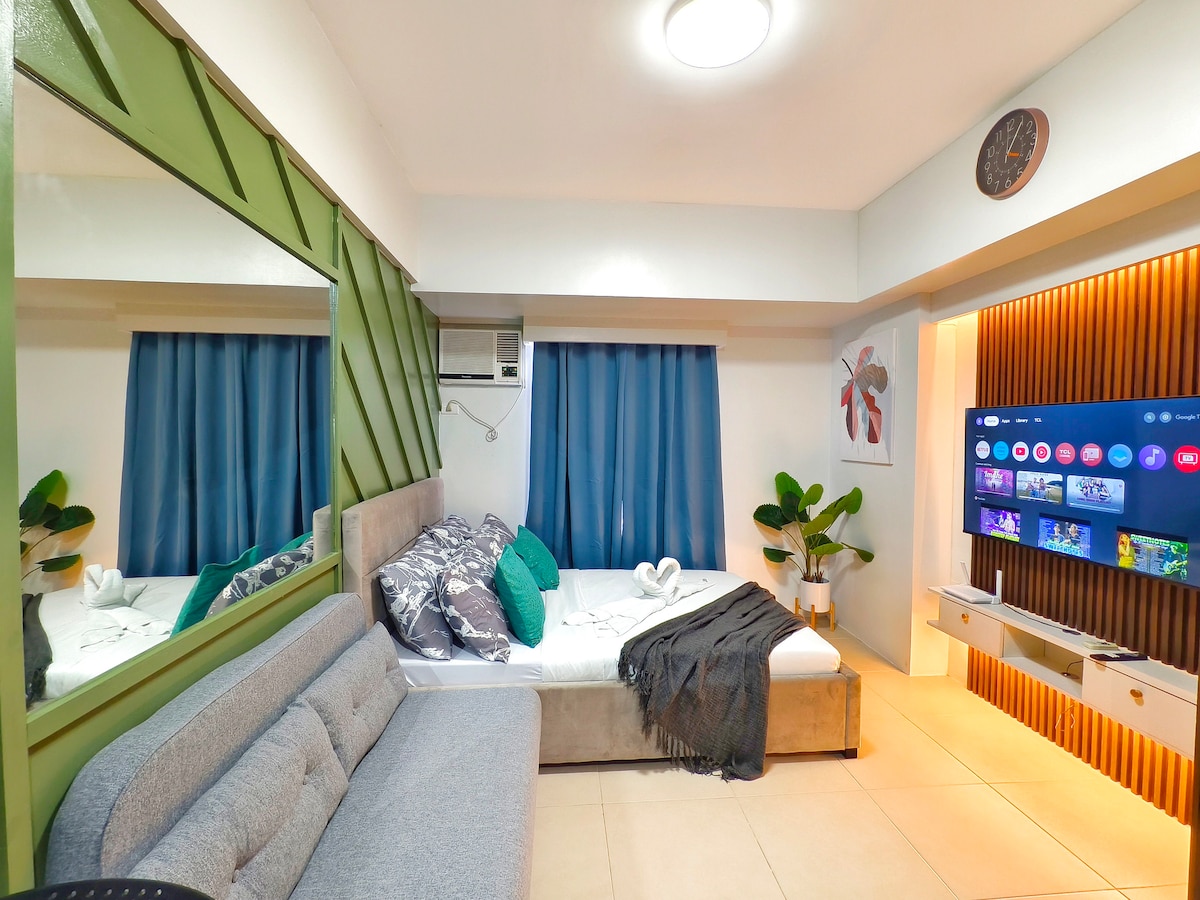 A cozy studio unit is shown, featuring a spacious bed adorned with decorative pillows and a soft blanket. A comfortable sofa is positioned beside a large mirror and distinct green accent wall. A wall-mounted smart TV displays various streaming options, while two large windows are dressed with blue curtains.
