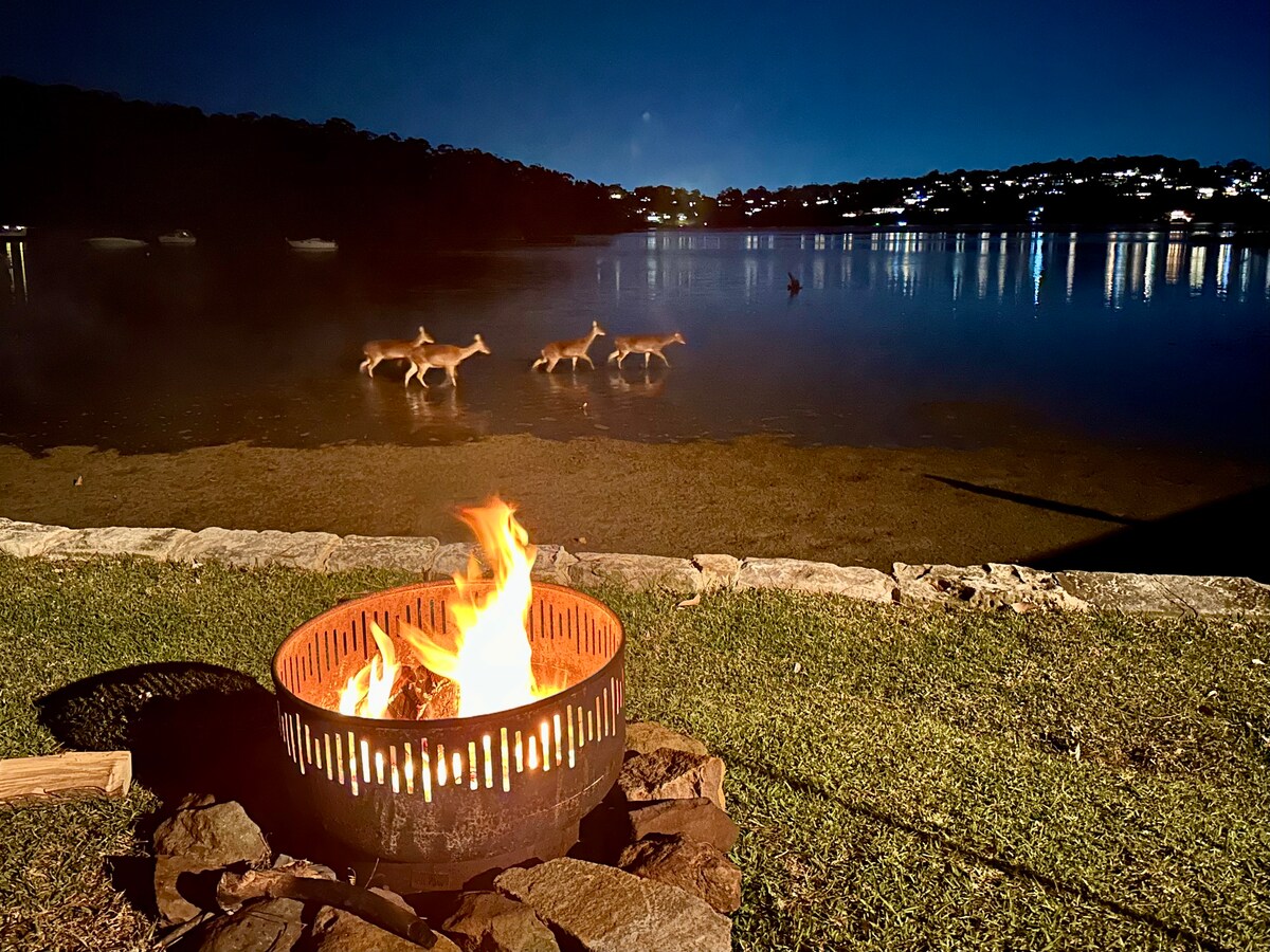 A tranquil evening scene features a fire pit with flames flickering in the foreground, surrounded by rocks. The serene waters reflect distant lights, while deer can be seen wading near the shoreline. The natural landscape is illuminated by soft moonlight.