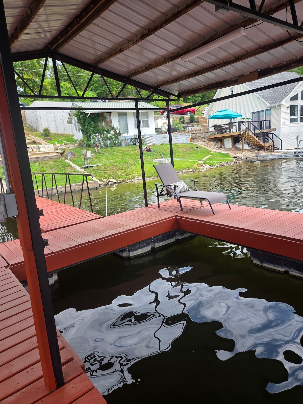 A private dock is depicted, featuring a wooden deck with a comfortable lounge chair positioned for relaxation. Clear water reflects the sky and surrounding greenery, creating a serene lakeside ambiance. Nearby, a cabin is visible in the background, enhancing the tranquil setting.