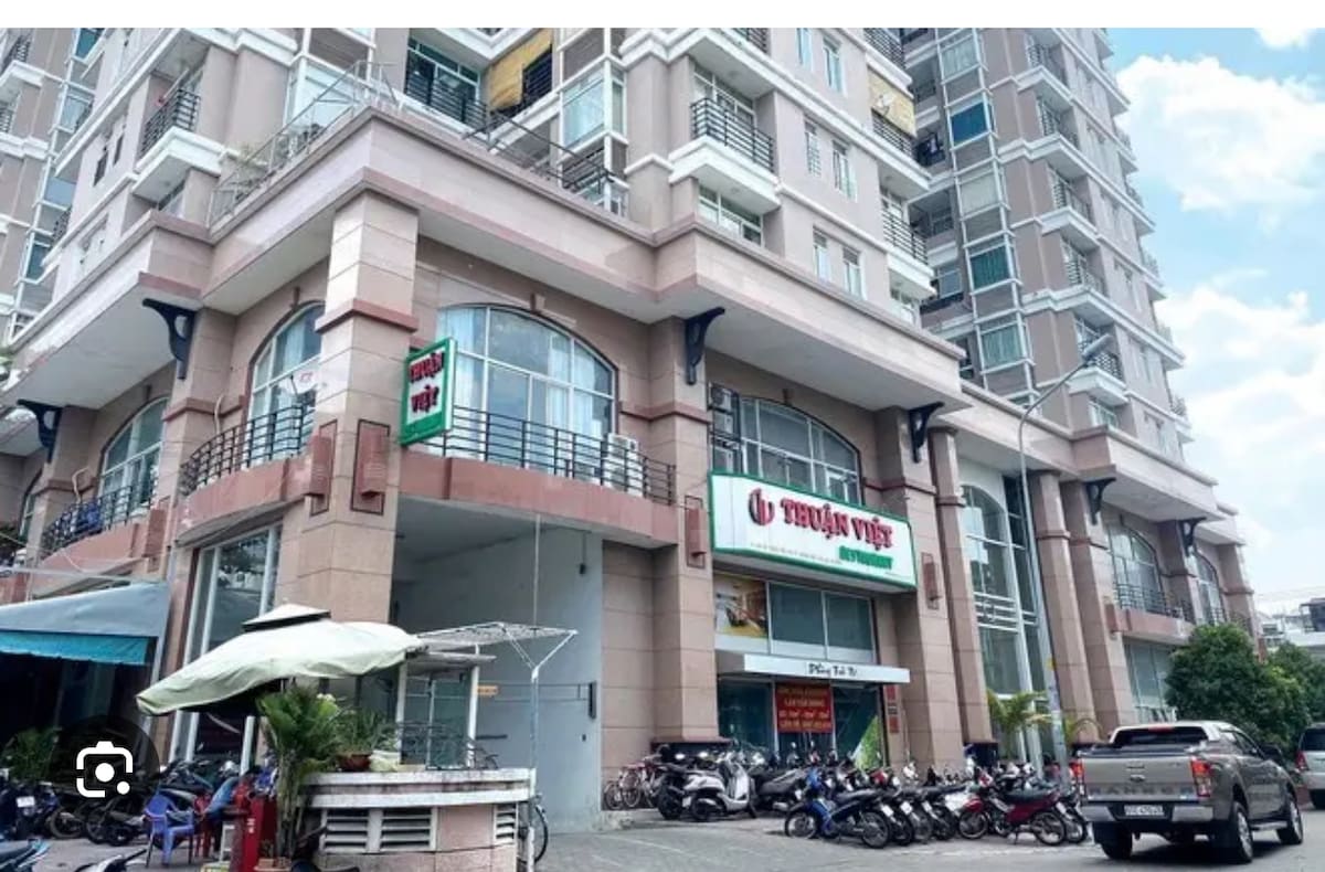 The exterior of a multi-story building is shown, featuring a prominent ground-level commercial space with signage. Vehicles are parked along the front, and an outdoor seating area is visible with greenery and shade. Clear skies are present above the structure.