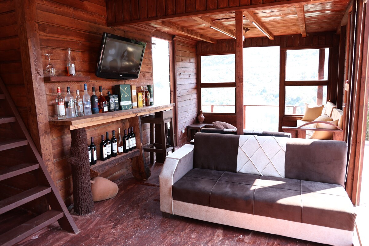 A cozy living area features a comfortable, modern sofa against a backdrop of wooden walls. A bar area is present with various bottles displayed on shelves. Natural light is let in through multiple large windows, providing views of the surrounding landscape.