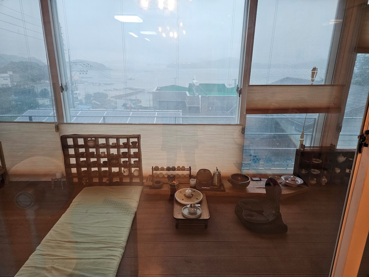 A serene indoor space is depicted, featuring a spacious mat on the floor and a variety of traditional tea utensils arranged on a wooden table. Large windows provide views of the tranquil surroundings, with a soft light emanating from overhead fixtures, enhancing the calming ambiance.