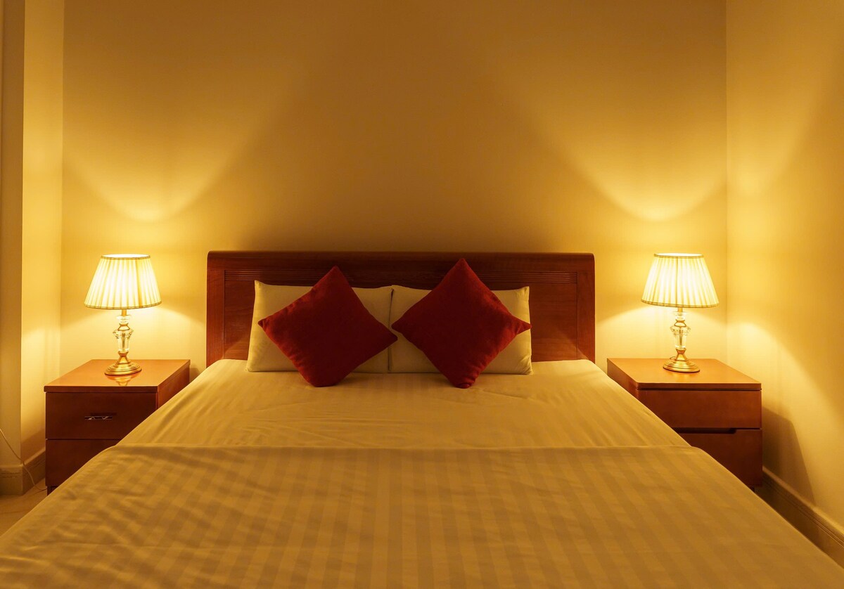 A well-lit bedroom features a spacious bed with white linens and decorative red accent pillows. Nightstands with stylish lamps sit on either side, casting a warm glow. Soft lighting creates a serene environment, ideal for relaxation.