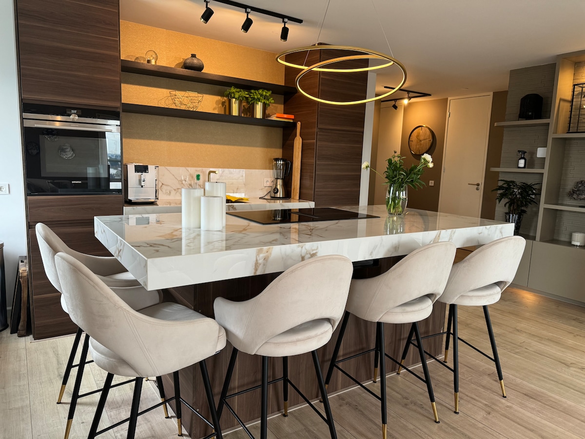 A modern kitchen space features a large marble island with soft, plush seating around it. The clean lines and warm wood accents are complemented by contemporary lighting above. Potted plants add a touch of greenery, and kitchen appliances are neatly integrated into the design.