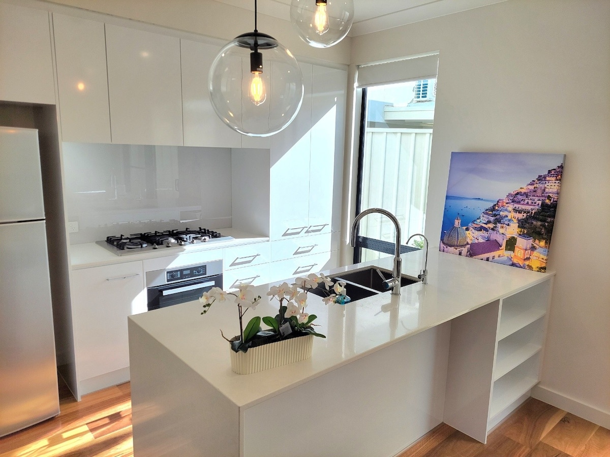 A modern kitchen features sleek white cabinetry and light wood flooring. An island with a built-in sink and a cooking area is highlighted by stylish pendant lighting. A vibrant piece of art depicting a coastal scene is displayed on the counter, adding a touch of color to the space.