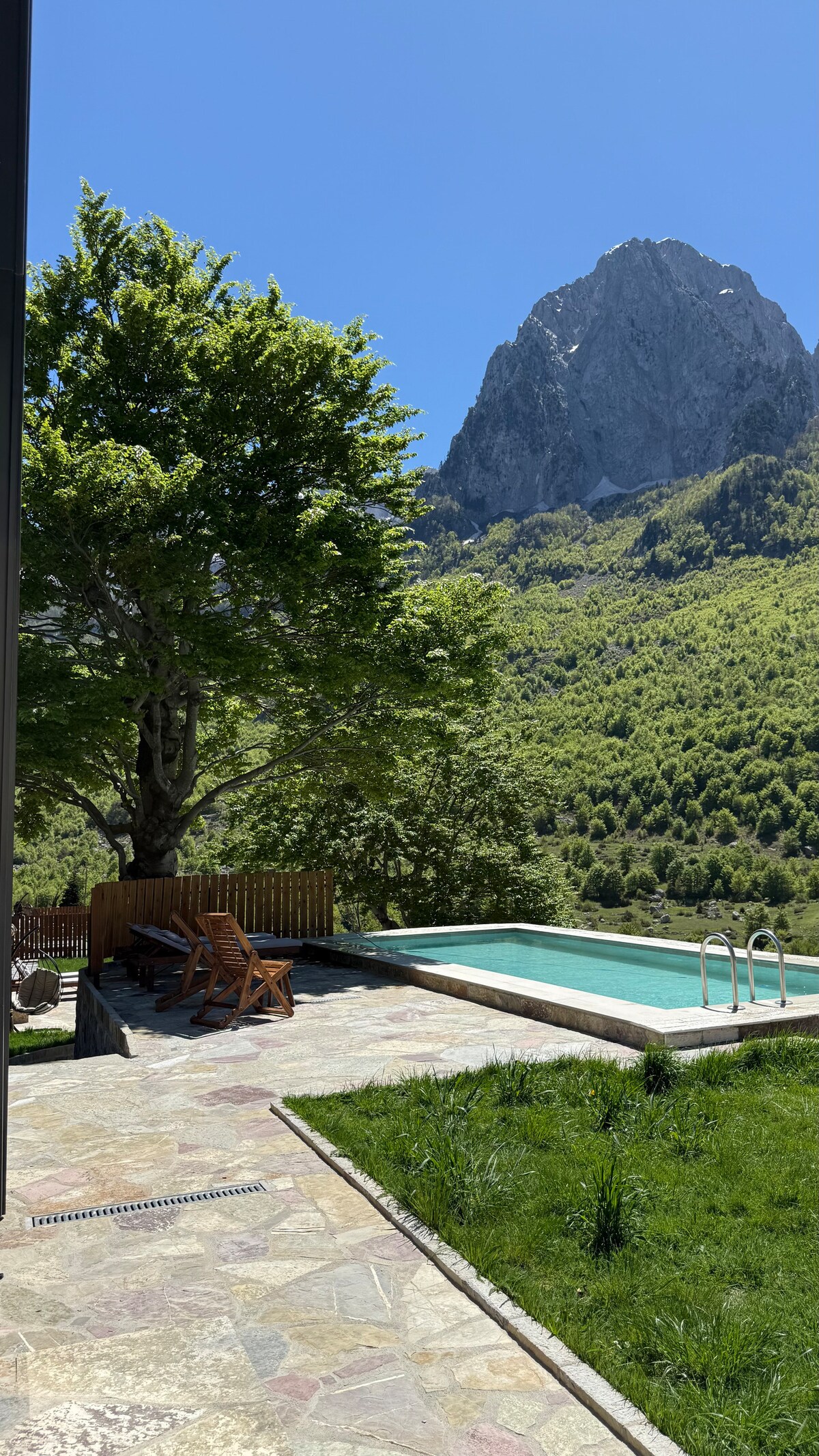 A serene outdoor area features a swimming pool surrounded by a stone patio. Wooden lounge chairs are positioned nearby, offering a space for relaxation. A large tree provides shade, while a majestic mountain looms in the background under a clear blue sky.