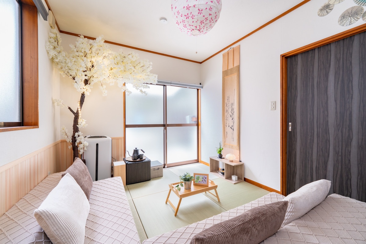 A serene Japanese-style room features two low sofas with soft cushions. A small wooden table sits in the center, accompanied by an electric kettle. A white cherry blossom tree decorates the space, and sliding glass doors provide natural light.