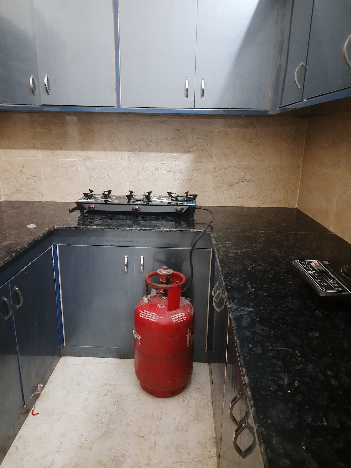 A compact kitchen space is equipped with a four-burner gas stove set against a marble backdrop. A red gas cylinder stands nearby, while dark cabinetry offers ample storage. Countertops feature a textured black surface, providing a practical work area for meal preparation.