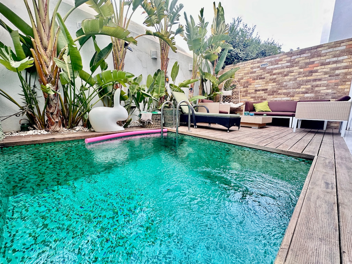 A private pool area is surrounded by tropical plants and features a sparkling turquoise pool. Lounge chairs are positioned next to the water, and a stone wall adds texture in the background. A pink pool float rests on the surface, inviting relaxation.