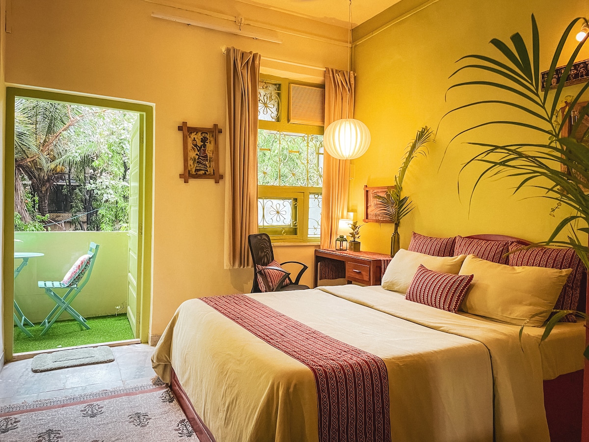 A brightly painted bedroom features a spacious bed with matching linens and decorative pillows. Natural light enters through large windows, illuminating a small balcony area with a bistro table and chairs. Indoor plants add a refreshing touch to the vibrant space.