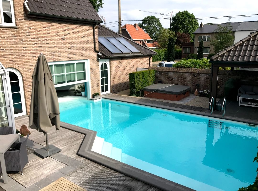 An outdoor area features a rectangular swimming pool bordered by light-toned decking. Comfortable seating is arranged nearby, including a large umbrella providing shade. A hot tub is located adjacent to the pool, with greenery and residential buildings visible in the background.