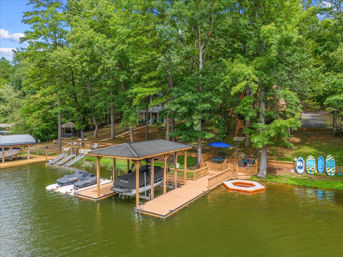 A private dock is seen on the lake, equipped with several covered boat slips. Surrounding trees provide shade, while a small deck and seating area are visible nearby. Watercraft are moored alongside the dock, enhancing the serene lakefront setting.