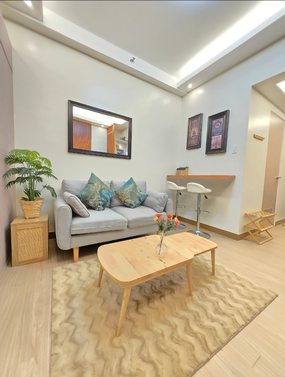 A cozy living area features a plush gray sofa adorned with patterned cushions. A small wooden coffee table sits atop a textured rug, with a vase of fresh flowers displayed. A mirror and wall art enhance the space, complemented by warm lighting.